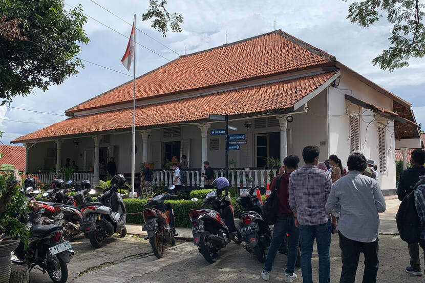 A group of people standing next to a historic center in Depok with scooters parked infront of the historic center.