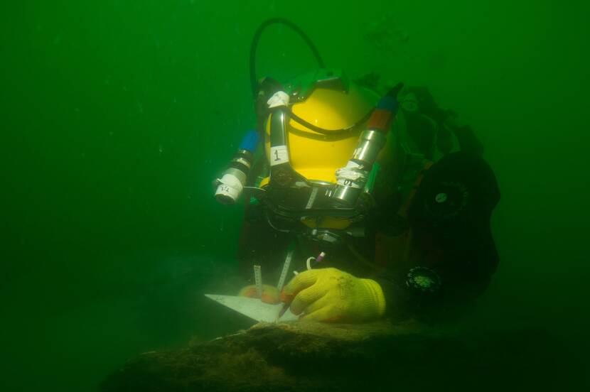 Diver holds writing block and pencil in hand while hovering over a wreck underwater