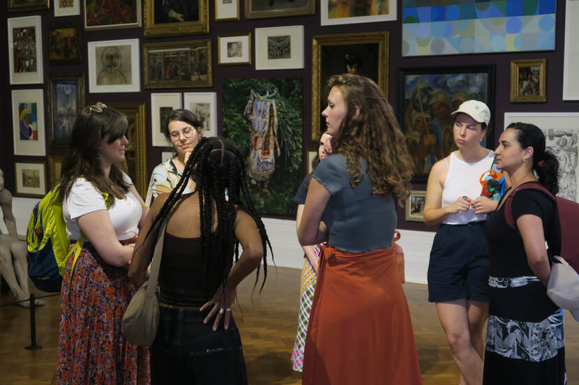 A circle of women with a wall full of framed artworks behind them.