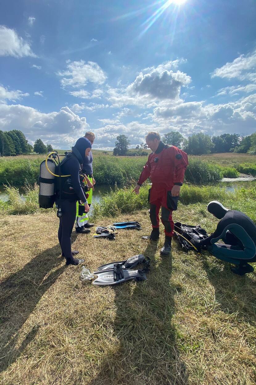 People in diving gear stand on the grass next to a river