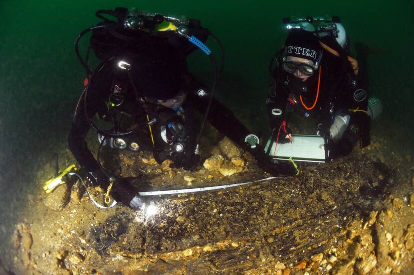 Two divers measure a wooden object on the bottom of the sea