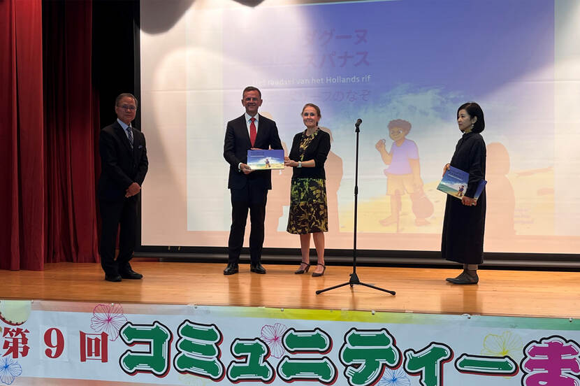 Standing on the left is Mr Ikehiro Michio. In the middle are Theo Peters and José Schreurs, with José Schreurs handing the children's book to Theo Peters while they also shake hands. On the right stands Kayoko Shimoji.